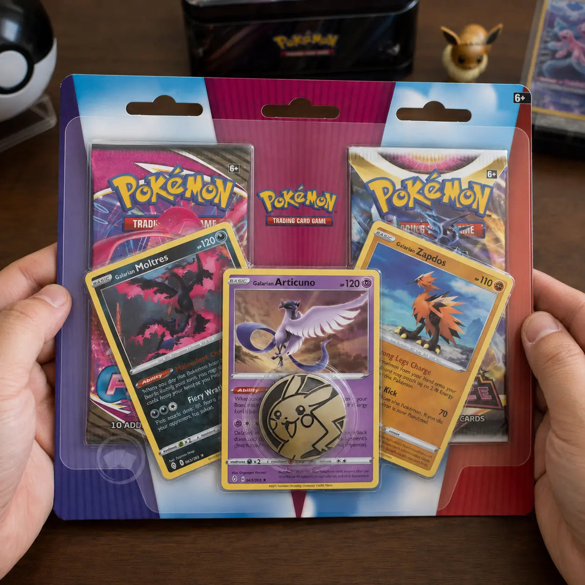 Hände halten Pokemon Galarian Birds Trio 2-Pack Blister auf Holztisch mit Evoli-Figur im Hintergrund